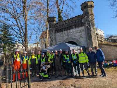 Vor dem steinernen Portal des Ernst-August-Stollens steht eine größere Gruppe von Personen in Signalwesten, Helmen und Schutzstiefeln in einer Reihe, dahinter ein aufgestelltes Zelt als temporäre Basisstation. Seitlich sind Bäume, Gebäude und Ausrüstung zu sehen, während ein Kinderwagen im Vordergrund auf die gemischte, interdisziplinäre Gruppe hindeutet. Die Szene dokumentiert die Einsatzbereitschaft des Messteams vor Beginn der Befahrung.  