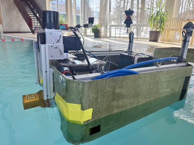 TB1 schräg von vorn im Schwimmbecken: Ein bootsähnlicher, rechteckiger Rumpf mit aufgesetzter Metallrahmen‑Konstruktion, Sensor oben, Kabel und blaue Schläuche im Inneren. Am hinteren Ende ragt auf einem Mast ein dunkles Prisma nach oben; im Hintergrund sind große Fensterfronten und Pflanzen des Freizeitbads zu sehen.