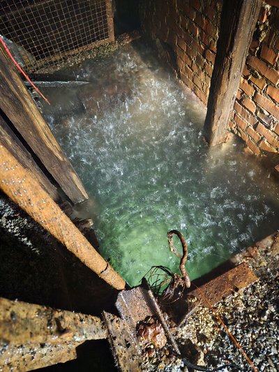 Blick von oben in eine von Ziegel- und Holzbalken begrenzte Schachtstruktur, in der türkisgrünes Wasser. Auf der Oberfläche sind die Einschläge der Regentropfen zu sehen. Rostige Metallteile, Kies und ein nach unten führendes Kabel deuten auf technische Installationen und Messaufbauten im Rahmen der Tiefensondierung hin. Das von unten Wasser ist Teil der hydrogeologischen Untersuchungen im Knesebeckschacht.  