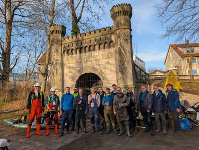 Eine Gruppe von etwa 15 Personen steht frontal vor dem historischen, burgähnlichen Portal des Ernst-August-Stollens im Freien, teils in Arbeitskleidung mit Helmen und Wathosen, teils in Freizeitkleidung. Im Vordergrund liegen Ausrüstung, Leitungen und ein SUP, die auf die untertägige Messkampagne und Tiefensondierungen im Knesebeckschacht hinweisen. Die Personen blicken lächelnd in die Kamera und markieren den erfolgreichen Abschluss des EAGruMo-Einsatzes.  