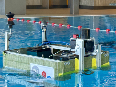 Seitliche Ansicht der schwimmenden Plattform TB1 in der Beckenmitte: Ein rechteckiger, grüner Rumpf mit aufgelegtem Technikmodul aus Metallprofilen, oben Sensorik und Kabel, seitlich ein aufgeklemmtes EAGruMo‑Logo. Im Hintergrund ziehen sich rote Schwimmleinen über die Wasseroberfläche des Hallenbads.