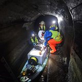 In einem engen, niedrigen Stollen arbeiten mehrere mit Helmen und Warnwesten ausgestattete Personen bei Stirnlampenlicht an einem mit Kisten, Kabeln und Messgeräten beladenen Stand-Up-Paddle-Board, das im flachen Wasser steht. Im Vordergrund liegt eine große Kunststoffkiste mit aufgewickeltem Kabel, die als mobile Plattform für die Messtechnik des EAGruMo-Einsatzes im Knesebeckschacht dient. Die dunklen, feuchten Stollenwände rahmen die Szene nah und seitlich ein.  
