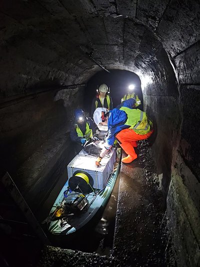 In einem engen, niedrigen Stollen arbeiten mehrere mit Helmen und Warnwesten ausgestattete Personen bei Stirnlampenlicht an einem mit Kisten, Kabeln und Messgeräten beladenen Stand-Up-Paddle-Board, das im flachen Wasser steht. Im Vordergrund liegt eine große Kunststoffkiste mit aufgewickeltem Kabel, die als mobile Plattform für die Messtechnik des EAGruMo-Einsatzes im Knesebeckschacht dient. Die dunklen, feuchten Stollenwände rahmen die Szene nah und seitlich ein.  