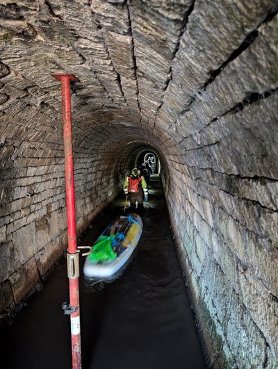 Frontaler Blick in einen langen, gemauerten Stollen mit flachem Wasser, in dem eine Person in Schutzkleidung und Wathose läuft und ein beladenes Stand-Up-Paddle-Board hinter sich herzieht. Im Hintergrund sind weitere Personen und die Öffnung des Stollens zu erkennen, während links im Vordergrund eine rote Stütze steht Die Szene zeigt den materialgestützten Transport im Rahmen der EAGruMo-Befahrung.
