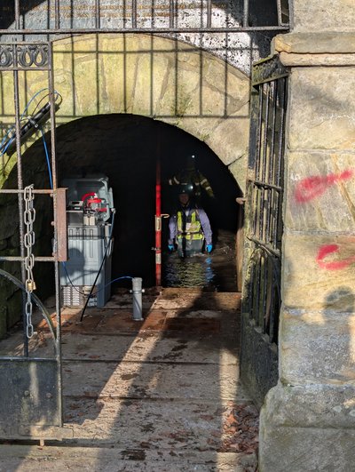 Durch das geöffnete Gittertor des Ernst-August-Stollens sind zwei Personen in Wathosen, Helmen und Warnwesten zu sehen, die kniehoch im Wasser im dunklen Stollen stehen. Links im Eingangsbereich steht ein graues Technikmodul mit Kabeln und Schläuchen, das Teil des Monitoring im EAGruMo-Projekts ist. Sonnenlicht fällt von außen auf den Boden und kontrastiert mit dem dunklen Stolleninneren.  